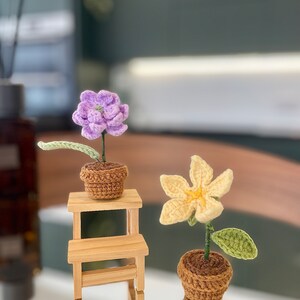 May include: Two crocheted flower plants in brown pots. The purple flower is on a small wooden shelf. The yellow flower is on the table below the shelf.