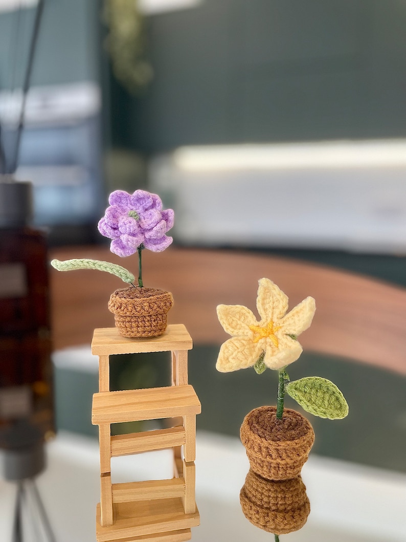 May include: Two crocheted flower plants in brown pots. The purple flower is on a small wooden shelf. The yellow flower is on the table below the shelf.
