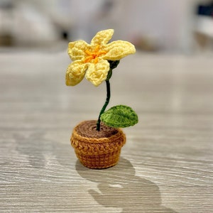 May include: A small crocheted flower in a yellow and orange colour scheme is potted in a brown crocheted pot. The flower has a single green leaf.