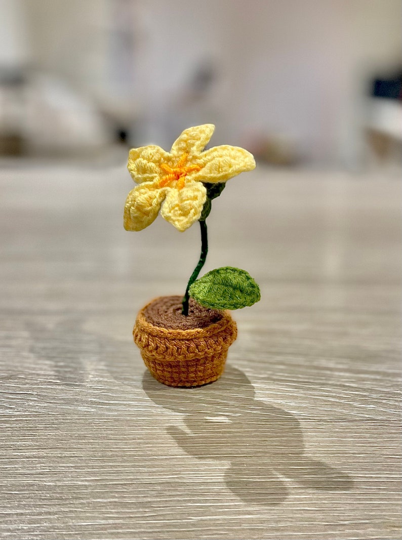 May include: A small crocheted flower in a yellow and orange colour scheme is potted in a brown crocheted pot. The flower has a single green leaf.
