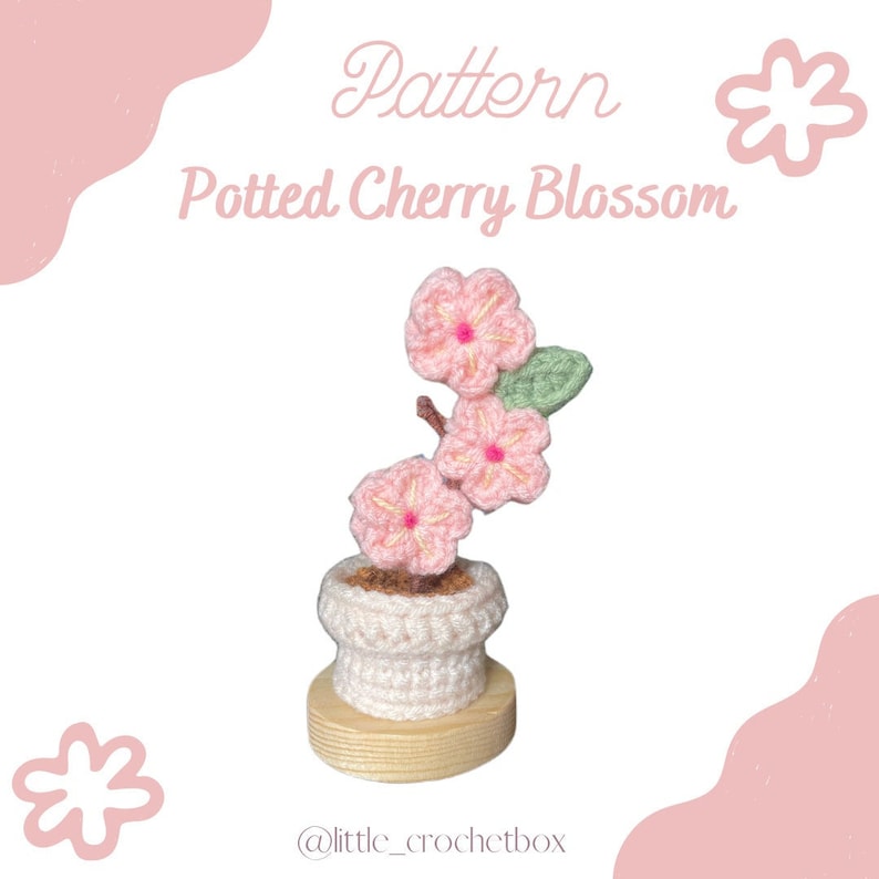K&ouml;nnte beinhalten: Geh&auml;kelte Topfpflanze mit Kirschbl&uuml;ten. Die Pflanze hat rosa Bl&uuml;ten mit gelben Zentren, gr&uuml;ne Bl&auml;tter und einen braunen Stiel, alles in einem wei&szlig;en Topf auf einem Holzsockel. Der Text "Pattern Potted Cherry Blossom" ist in Rosa dargestellt.