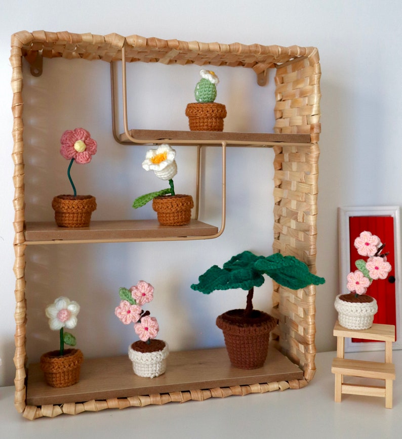 May include: A set of crocheted flower and plant pots displayed on a wooden shelf with a woven wicker frame. The shelf has three tiers and features a variety of colorful flowers and plants, including a cactus, a pink flower, a white flower, and a green plant.