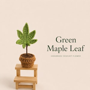 Pode incluir: Uma folha de ácer de croché verde presa a um caule castanho, plantada num vaso de croché castanho sobre um pequeno banco de madeira. O texto Green Maple Leaf HANDMADE CROCHET FLOWER está impresso à direita em letras verde-escuras.