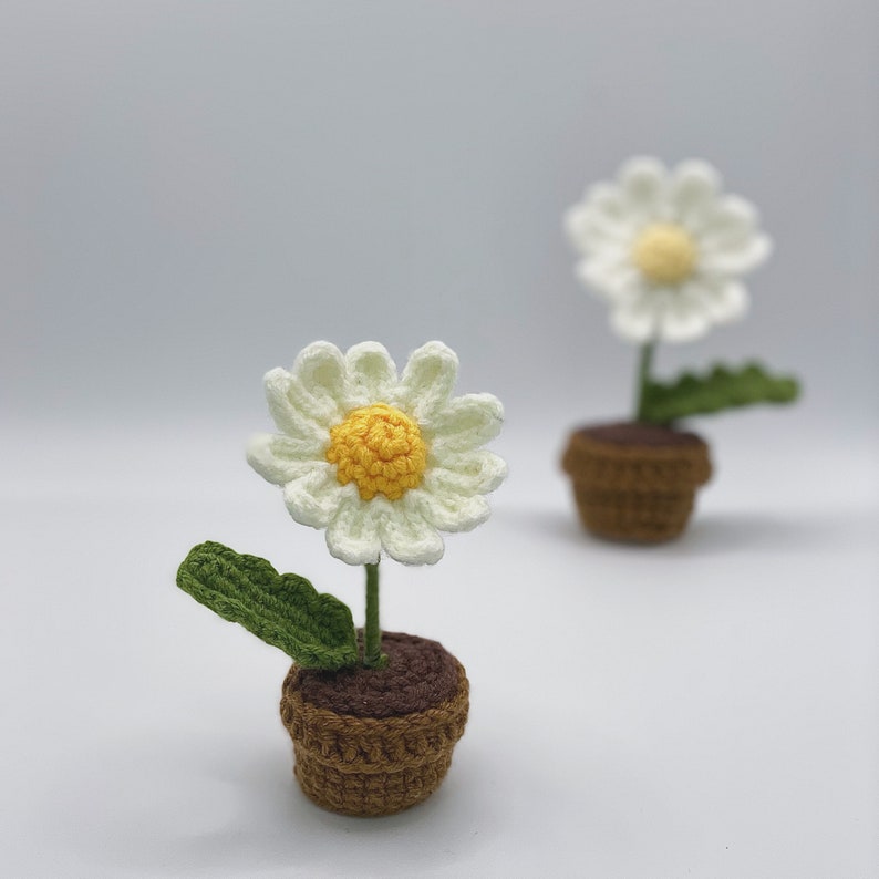 May include: Two crocheted daisy flowers in brown pots. The daisies have white petals and yellow centers. The pots are brown and have a textured surface.