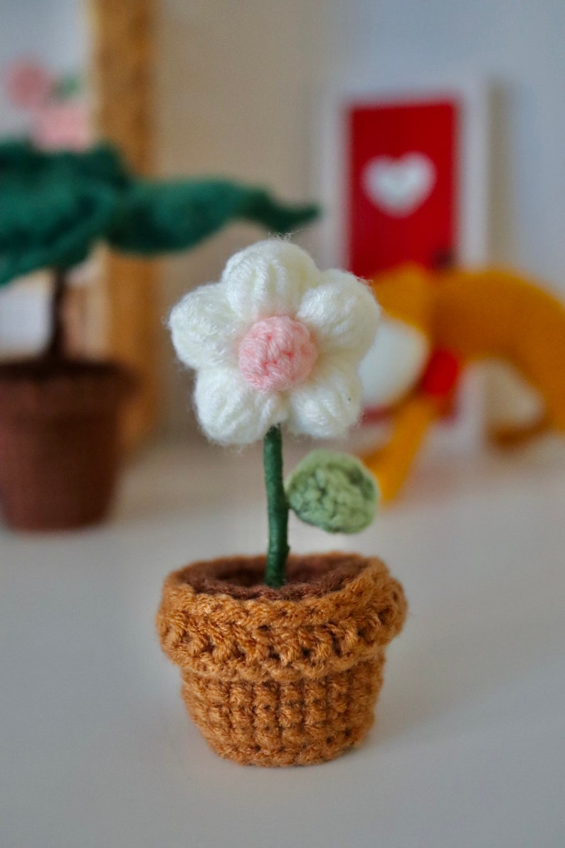 May include: A small crocheted flower in a brown pot. The flower is white with a pink center and has a green leaf.