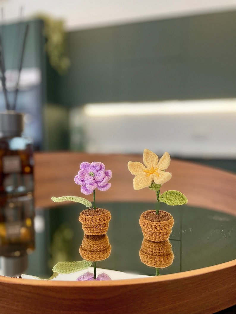 May include: Two crocheted flower pots, one with a purple flower and the other with a yellow flower, are displayed on a wooden tray with a mirrored surface.