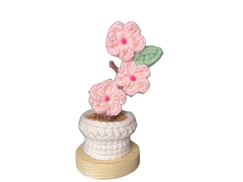 K&ouml;nnte beinhalten: Handgefertigte H&auml;kelblumenanordnung mit rosa Bl&uuml;ten und gelben Zentren sowie gr&uuml;nen Bl&auml;ttern. Die Blumen befinden sich in einem wei&szlig;en H&auml;keltopf auf einem Holzsockel und bilden ein dekoratives St&uuml;ck.