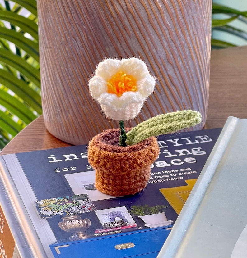 May include: A crocheted flower in a brown pot with a green leaf. The flower is white with a yellow center. The pot is sitting on a book with the text "Style Living Space".