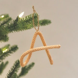 Peut inclure: Une décoration en forme de lettre A tricotée de couleur pêche clair est suspendue à un sapin de Noël artificiel vert avec des lumières blanches.