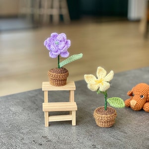 May include: Two crocheted flower plants in brown pots. The purple flower is on a small wooden stool. The yellow flower is on the table next to a crocheted brown dog.
