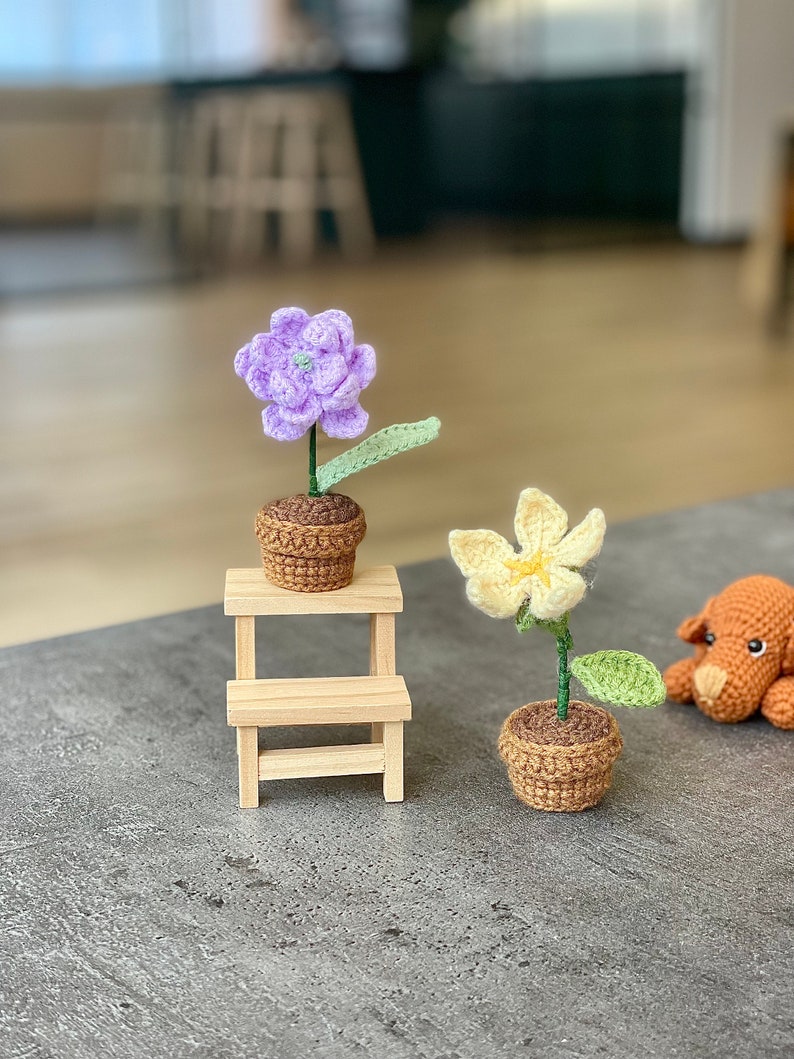 May include: Two crocheted flower plants in brown pots. The purple flower is on a small wooden stool. The yellow flower is on the table next to a crocheted brown dog.