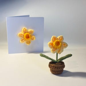 May include: A crocheted daffodil in a brown pot with green leaves. A matching daffodil is attached to a white card. The flower has yellow petals and a yellow center. The card is blank.