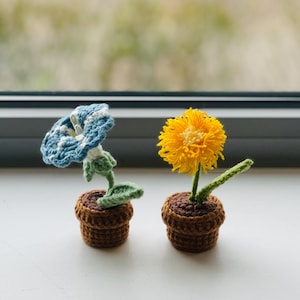 May include: Two crocheted potted plants. The plant on the left has blue and white flowers and green leaves. The plant on the right has yellow flowers and green leaves. Both plants are in brown pots.