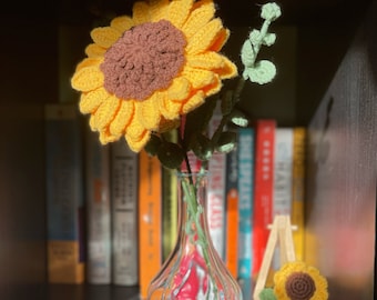 May include: A crocheted sunflower with brown and yellow petals in a clear glass vase on a bookshelf with colorful books.
