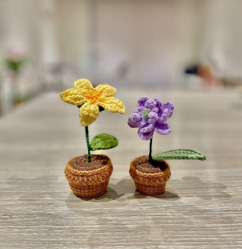 May include: Two crocheted potted plants, one with a yellow flower and one with a purple flower. The pots are brown and the plants have green leaves.