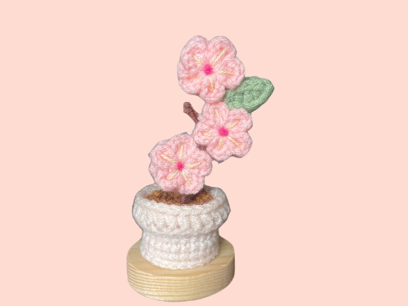 K&ouml;nnte beinhalten: Handgefertigtes H&auml;kelblumenarrangement mit rosa Bl&uuml;ten mit gelben Zentren und gr&uuml;nen Bl&auml;ttern. Die Blumen befinden sich in einem wei&szlig;en H&auml;keltopf mit Holzsockel, vor einem zartrosa Hintergrund. Ein dekorativer Artikel.