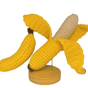 May include: A crocheted banana with a yellow peel and a cream-colored inside. The banana is displayed on a wooden stand.