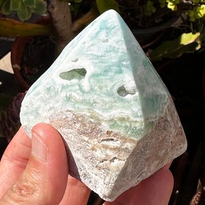 May include: A green and white pyramid-shaped crystal with a rough, natural texture. The crystal has a layered appearance with a light green color in the center and a white and brown color on the outside.