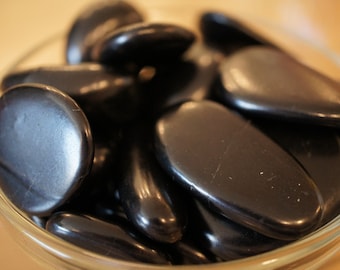 May include: A glass bowl filled with smooth, black polished stones. The stones are various sizes and shapes, and they have a glossy finish.