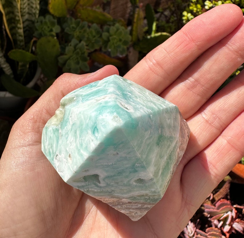 May include: A green and white, faceted, polished,  crystal gemstone. The crystal is shaped like a pyramid with a rounded top.