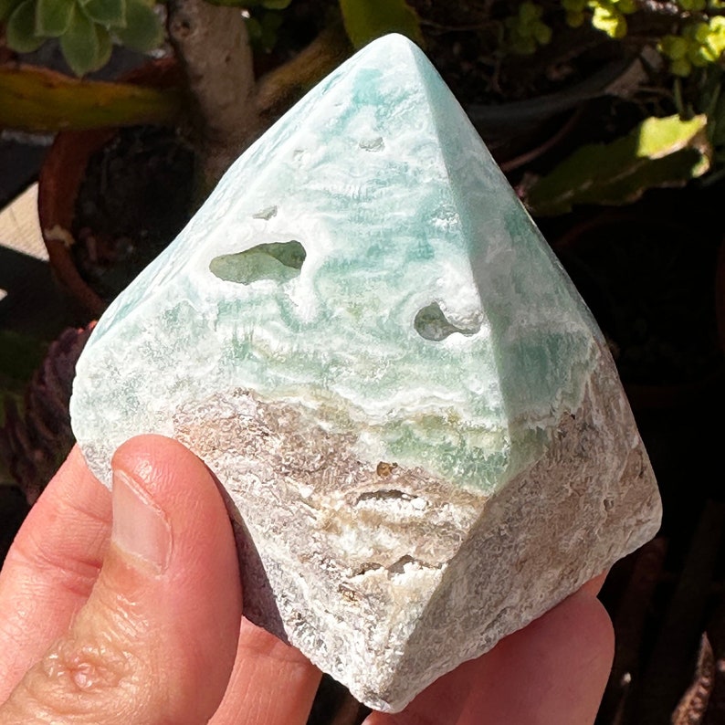 May include: A green and white pyramid-shaped crystal with a rough, natural texture. The crystal has a layered appearance with a light green color in the center and a white and brown color on the outside.