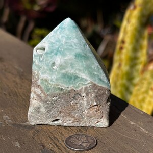 May include: A green and brown celestite crystal point, also known as a celestite tower, with a rough, natural finish. The crystal is about 3 inches tall and is sitting on a wooden surface.