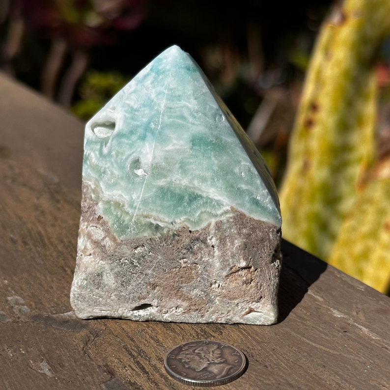 May include: A green and brown celestite crystal point, also known as a celestite tower, with a rough, natural finish. The crystal is about 3 inches tall and is sitting on a wooden surface.