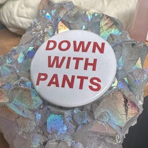 May include: A white button with red text that reads "DOWN WITH PANTS" on a bed of iridescent crystals.