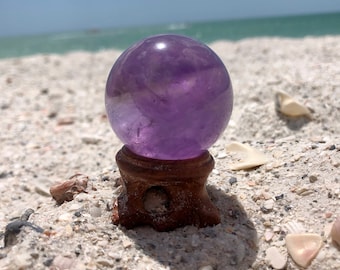 Op de afbeelding: Een grote, paarse amethist kristallen bol staat op een houten standaard. De bol is glad en glanzend, en de standaard is donkerbruin. De bol staat op een wit zandstrand.