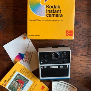 Puede incluir: Una cámara instantánea Kodak EK6 vintage con un cuerpo blanco y azul, una lente negra y un flash. La cámara está sobre una superficie de madera. La caja de la cámara es amarilla con un diseño de arcoíris y el texto "Kodak instant camera" y "Uses KODAK Instant Print Film PR 10".
