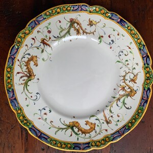 May include: A white ceramic plate with a scalloped edge and a colorful floral design featuring seahorses and scrolls. The plate has a blue, green, yellow, and gold border.