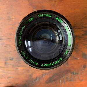 May include: A black camera lens with green text that reads "QUANTA RAY AUTO ZOOM 75-200mm 1:4.5 MACRO 855 MC".