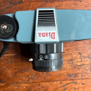 May include: A blue and black Diana camera with a lens that has a black and white striped ring around it. The lens has a small knob on the side and a "B" and "O" setting.
