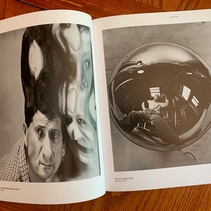 May include: A black and white photograph of a man with a distorted reflection of his face above his head. The reflection is created by a curved mirror. The man is looking at the camera with a serious expression. The photograph is titled "Self Portrait with Model's Distorted Mirror".