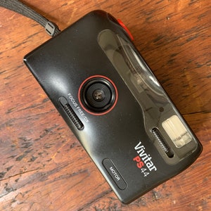 Puede incluir: Una cámara de película Vivitar PS 44 negra con una lente de enfoque libre roja y un flash incorporado. La cámara tiene una correa negra.