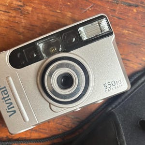 May include: Silver Vivitar 550PZ camera with a built-in flash and lens. The camera has a black lens ring and the words "Vivitar Series" are printed on the lens. The camera is on a wooden surface.