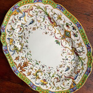 May include: A white oval ceramic serving platter with a colorful floral design. The border features green leaves, yellow dots, and blue and pink stripes. The center of the platter is decorated with a whimsical floral pattern in shades of yellow, green, blue, red, and orange.