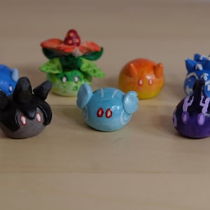 Puede incluir: Siete figuritas de arcilla hechas a mano, cada una representando una criatura diferente. Las criaturas son de varios colores, incluyendo azul, verde, naranja, morado y negro. Cada figurita tiene un diseño único y presenta detalles pintados.