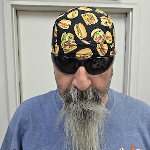 May include: A black skull cap featuring a repeating pattern of various cheeseburgers and hot dogs. The headwear is made of fabric and covers the top of the head, sitting just above the eyebrows. The print includes detailed illustrations of food.