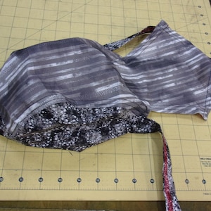 May include: A handmade head covering with a gray and white striped pattern on the main body and a speckled black and white pattern on the lower edge. The ties are a mix of red, white, and black.