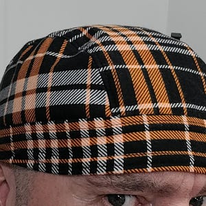 May include: A black, orange, and white plaid fabric hat with a brim. The hat is worn on a person's head.