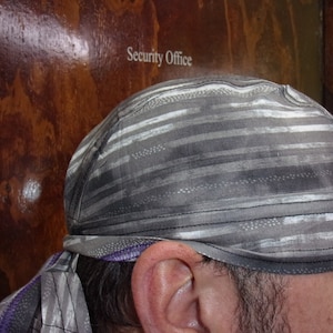 May include: A grey and white striped durag featuring a horizontal pattern with subtle black accents and a purple trim detail along the bottom edge. The fabric has a textured appearance with visible stitching along the seams and tie-back ends.