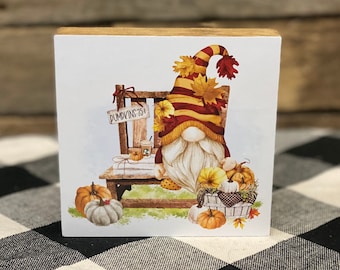 Könnte beinhalten: Ein Holzblock mit einem Gnom, der auf einem Stuhl sitzt und ein Schild mit der Aufschrift "Pumpkin 75¢" hält. Der Gnom trägt einen roten und gelben Hut mit Herbstblättern. Um den Gnom herum liegen Kürbisse und Kürbisse.