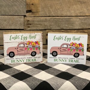 May include: Two Easter-themed wooden signs. Each sign features a pink truck carrying Easter eggs, with the text "Easter Egg Hunt" and "Bunny Trail." The signs also include text about balloons, arts & crafts, market, and bake sale.