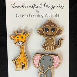 May include: Handcrafted wooden magnets featuring a giraffe, monkey, and elephant. The giraffe is yellow and orange, the monkey is brown, and the elephant is gray with pink and white accents. The magnets are on a white card with the text "Handcrafted Magnets by Gena's Country Accents."
