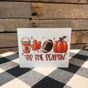 May include: A white card with a fall-themed illustration of a pumpkin, football, maple leaf, and a cup of coffee with whipped cream. The text "'Tis the season" is written in black below the illustration.