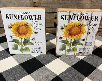 May include: Two wooden signs with a white background and black text that reads "ORGANIC SUNFLOWER Farm". Each sign features a yellow sunflower with green leaves and brown stems.  There are small black and yellow bees flying around the sunflowers. The text "FRESH PICKED" is below the sunflower and the text "SEEDS & FLOWERS FOR SALE" is below that. The signs are sitting on a black and white checkered surface.