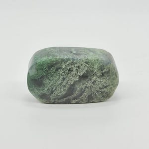 May include: A polished, rounded stone featuring a marbled pattern of deep forest green, sage, and charcoal gray. The surface displays intricate, moss-like inclusions and translucent patches, creating a smooth, organic texture throughout the piece.