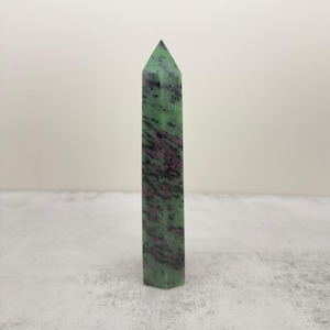 May include: A polished crystal tower with a hexagonal base and a pointed top. The stone features a mottled pattern of deep forest green and dark magenta or reddish-purple inclusions throughout the smooth, vertical, and cylindrical surface.