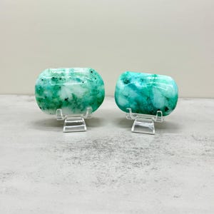 May include: Two polished, rectangular-shaped stones with rounded edges, featuring a marbled pattern of vibrant green and white with small dark speckles. Each stone is displayed upright on a small, clear acrylic stand for support.